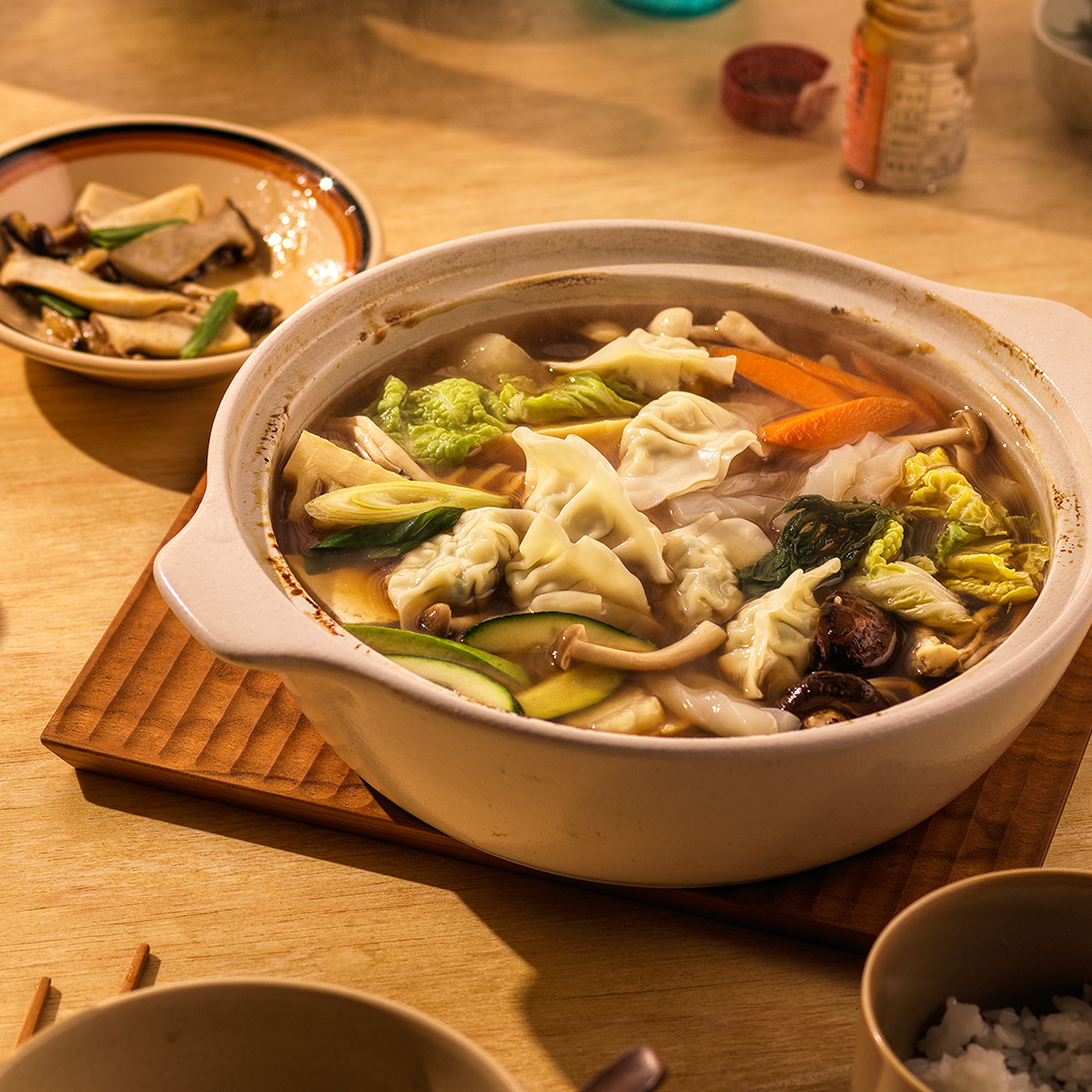 A warm bowl of gyoza soup with bibigo gyoza dumplings, tofu, sliced mushrooms, Chinese cabbage, zucchini, and carrots, all in a light, savory broth.