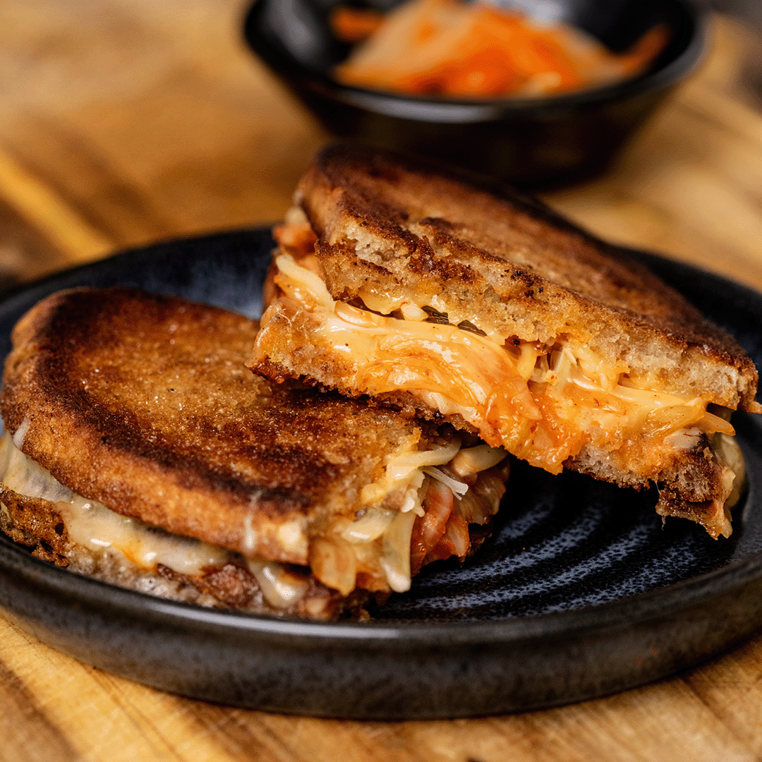 A close-up of a sliced bibigo kimchi cheese sandwich, revealing its gooey melted cheese, vibrant red kimchi, and crispy golden-brown bread, perfect for a savoury and tangy bite.