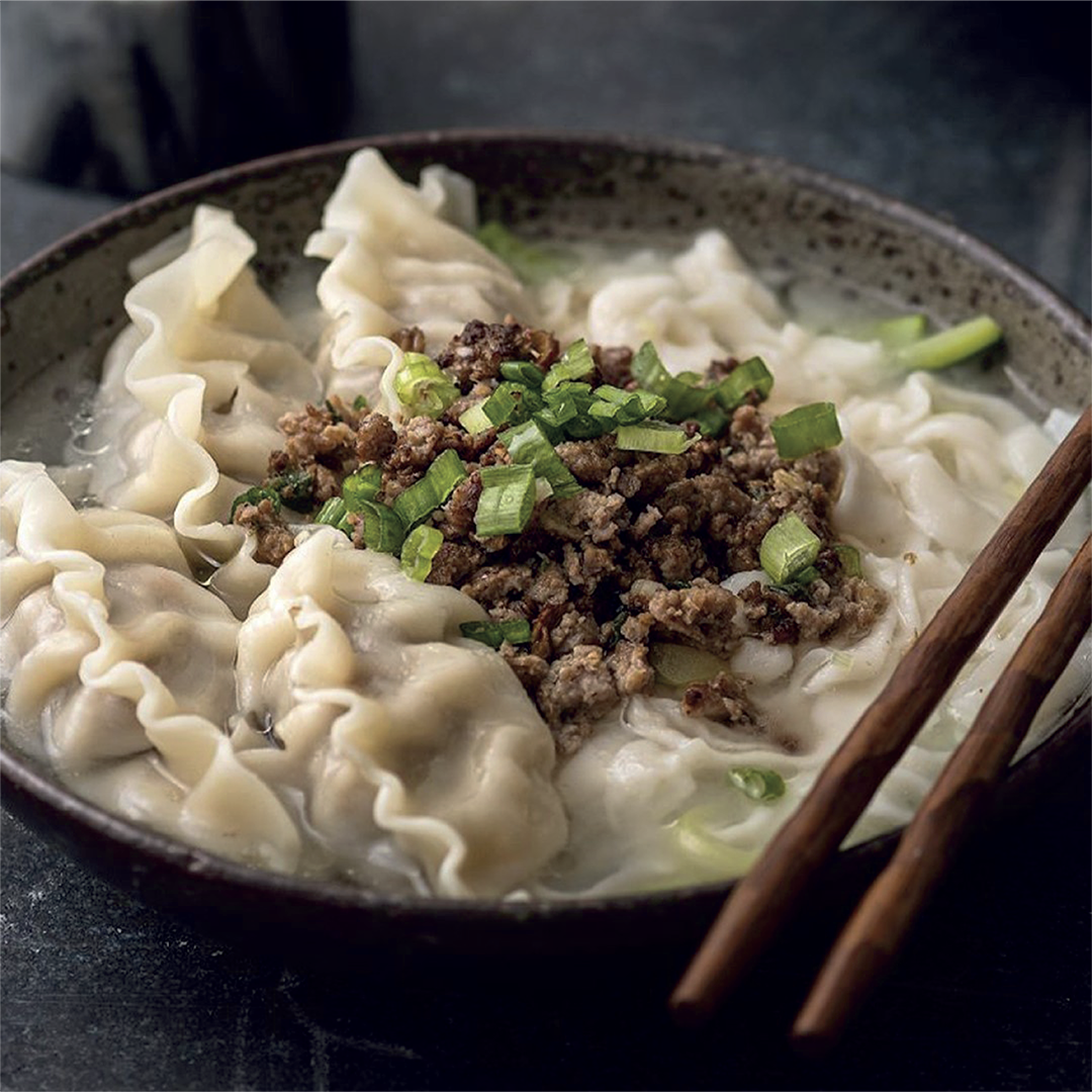 A bowl of bibigo Mandu Kal-Guksu, featuring hearty Korean noodles in a savory broth with dumplings, minced beef, and green onions on top.