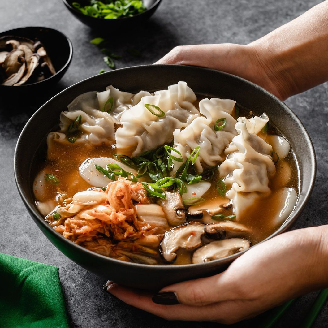 A bowl of Tteok Mandu Guk, combining bibigo Plant-based Kimchi Flavoured Mandu, Tteok rice cakes, shiitake mushrooms, and kimchi in a clear broth. The dish is garnished with green onions.