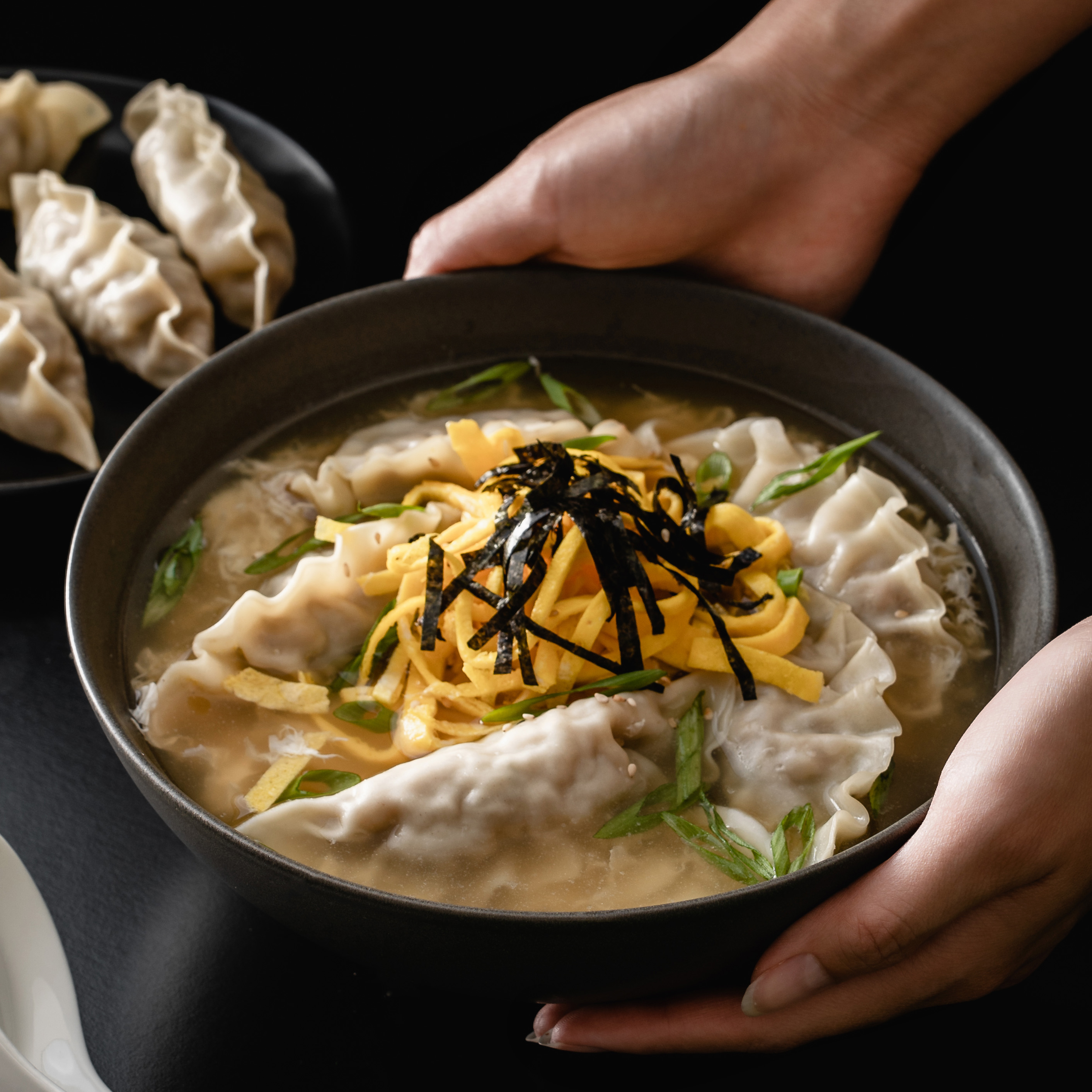 A bowl of Mandu Guk with bibigo Mandu, garnished with thin egg yolk strips, seaweed, sesame oil, and spring onions. The savory broth is filled with dumplings, egg strands, and a rich blend of flavors.